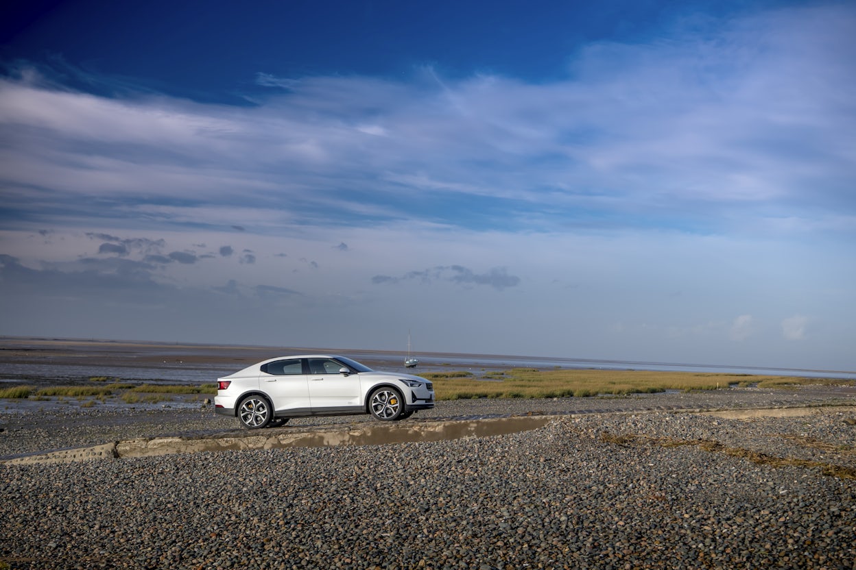 A white Polestar 2 on a stone beach under a blue sky.