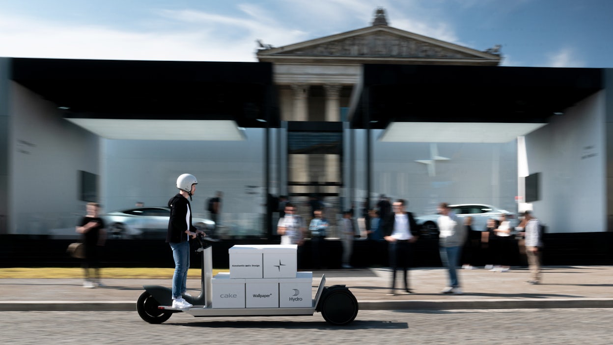 Man riding a scooter with boxes attached to it, with a blurry Polestar space in the background.