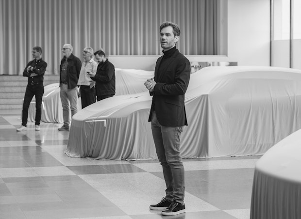 A man stands in the foreground, speaking, with four people in the background. Several vehicles covered with fabric are lined up behind them in a showroom-like setting.