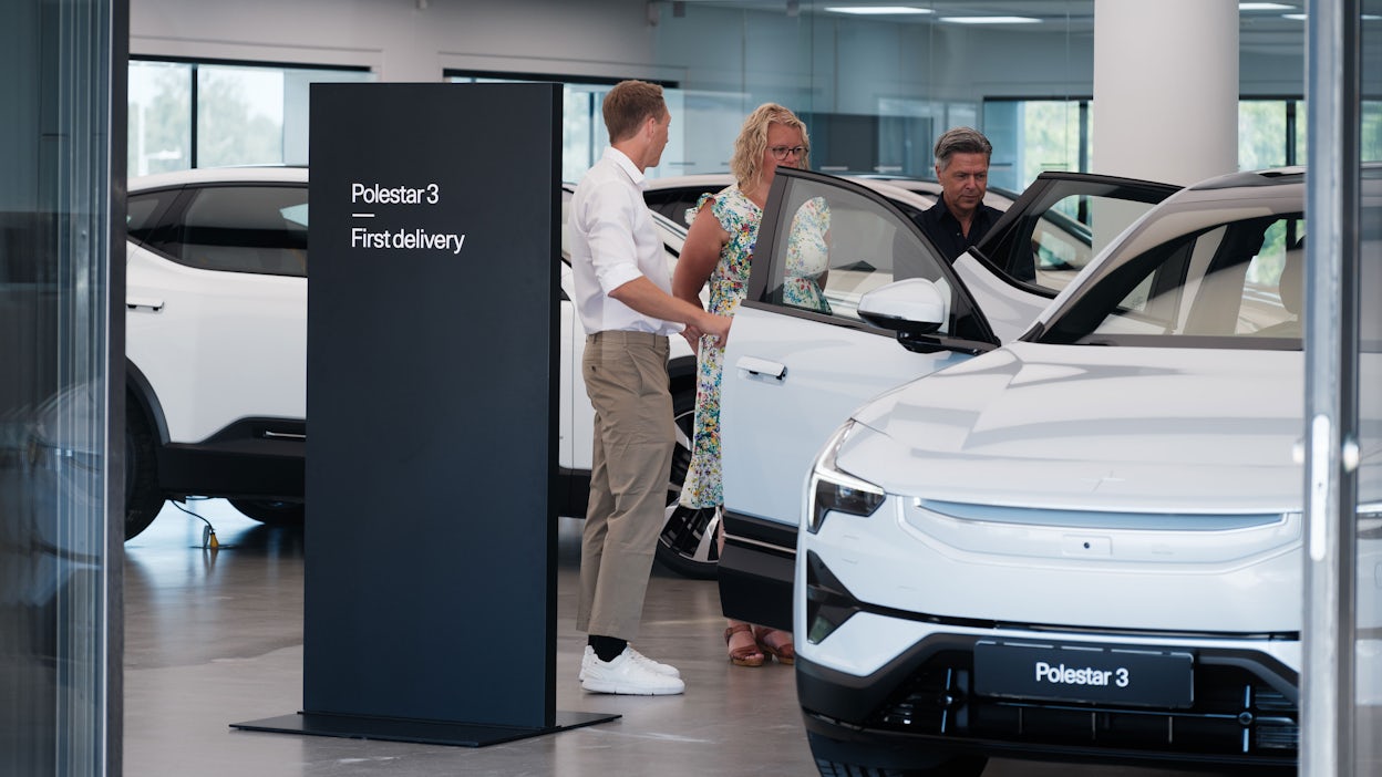 Inside Polestar HQ with three people who have opened the doors of a Polestar 3 and are looking inside, with a sign reading "Polestar 3 - first delivery" to the left